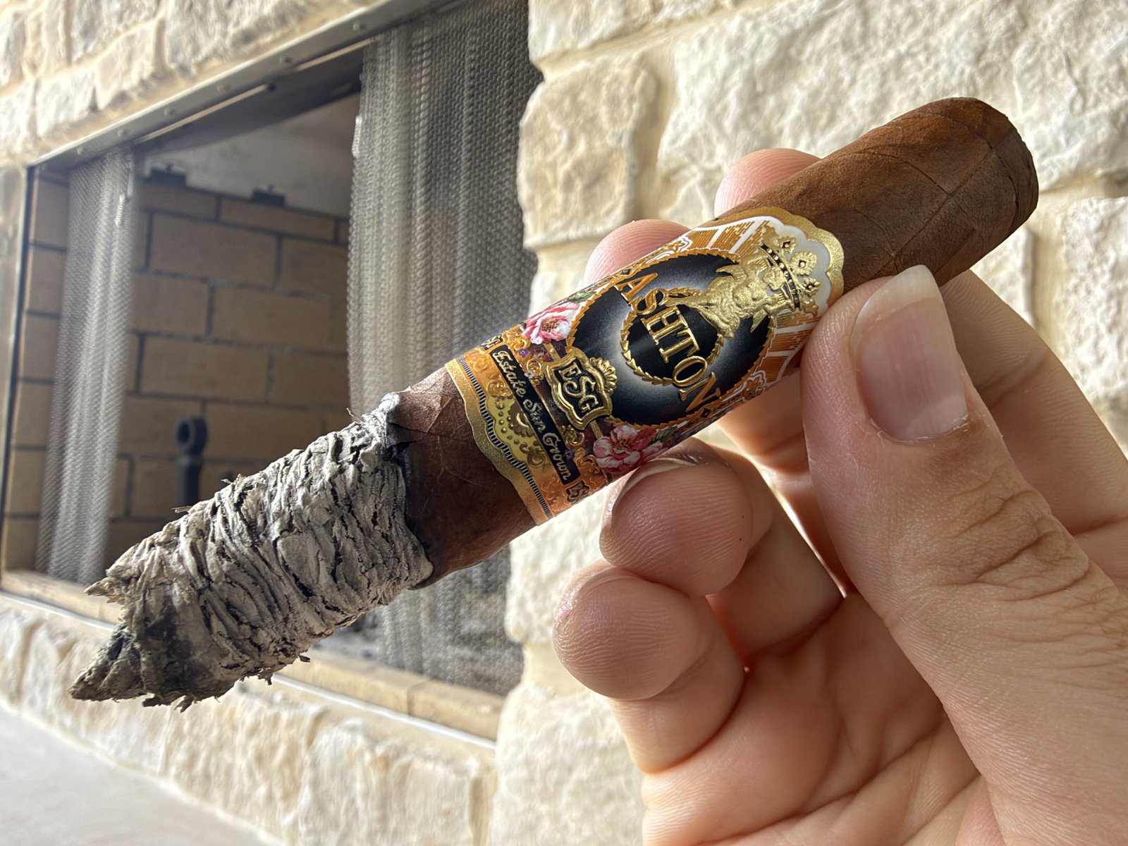 Ashton ESG 23 Year Salute Toro at the end of the second third with a long, slightly flaky gray ash stack and a wavy burn line, held in hand in front of a stone fireplace