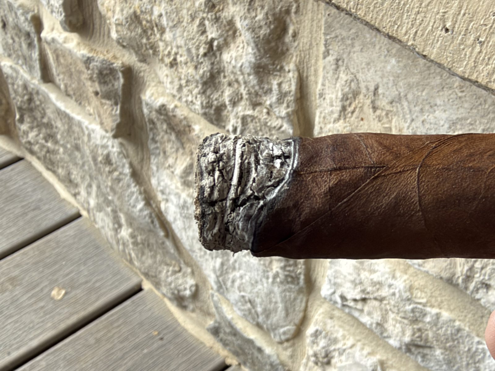 Ashton VSG Torpedo about half an inch in, with a short gray ash and a burn line running visibly crooked from one side to the other, in front of a stone wall