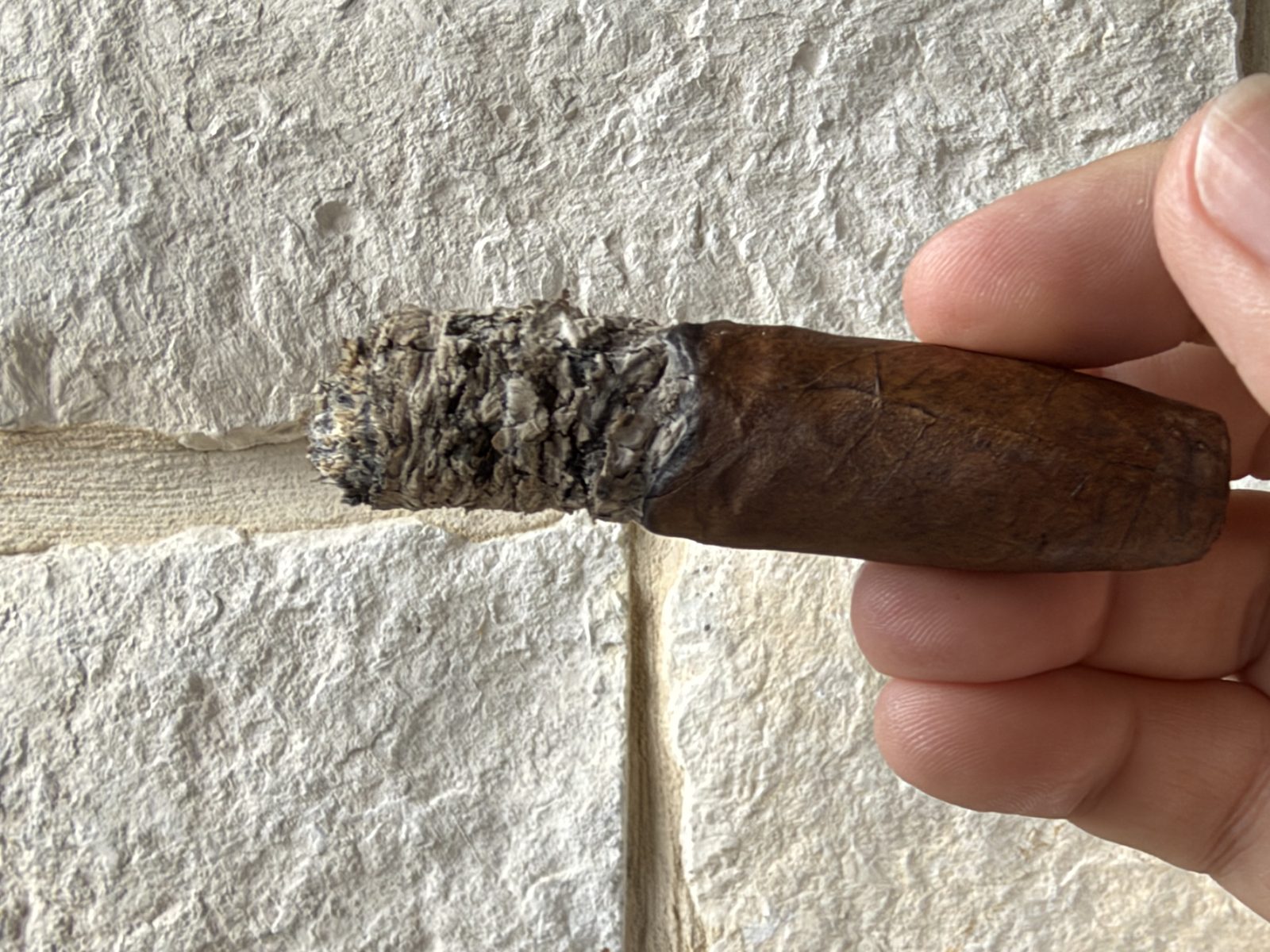 Ashton VSG Torpedo in the final third with the band removed, a short ragged gray ash and a fairly even burn line, held in fingers in front of a stone wall