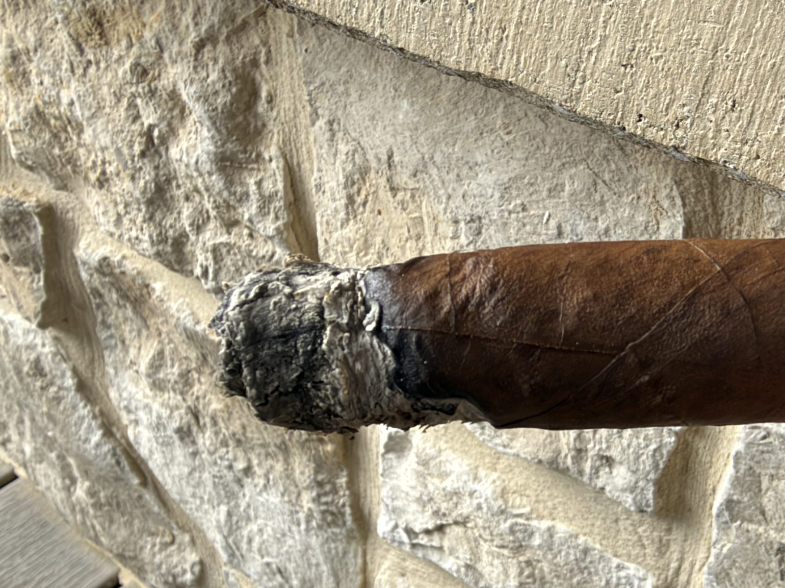 Ashton VSG Torpedo midway through the second third, ash knocked back to show an uneven, charred burn line with one section lagging behind the rest, in front of a stone wall