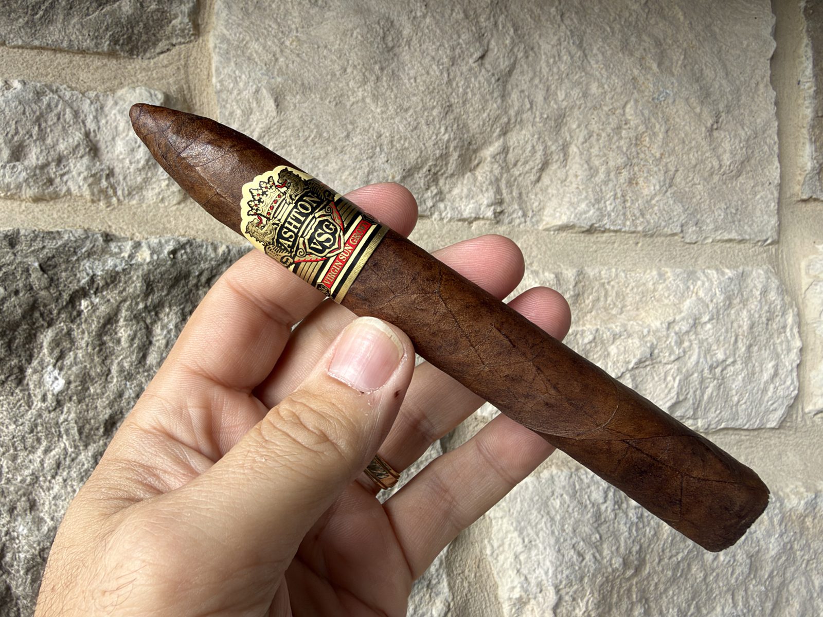 Unlit Ashton VSG Torpedo held in hand against a stone wall, dark reddish-brown Ecuadorian Sumatra wrapper with the gold, black and red Ashton VSG band