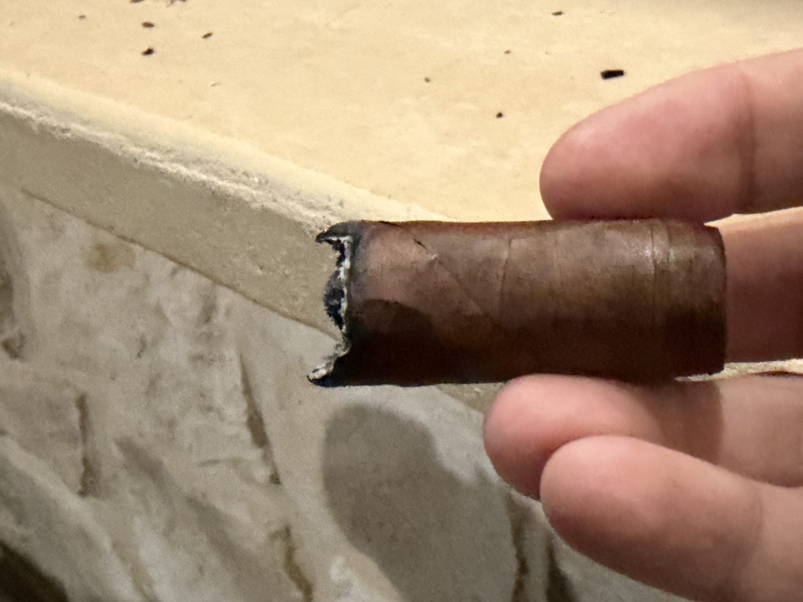 Casa Magna Colorado Robusto in the final third with the band removed, short and uneven burn edge at the foot, held in fingertips over a stone ledge