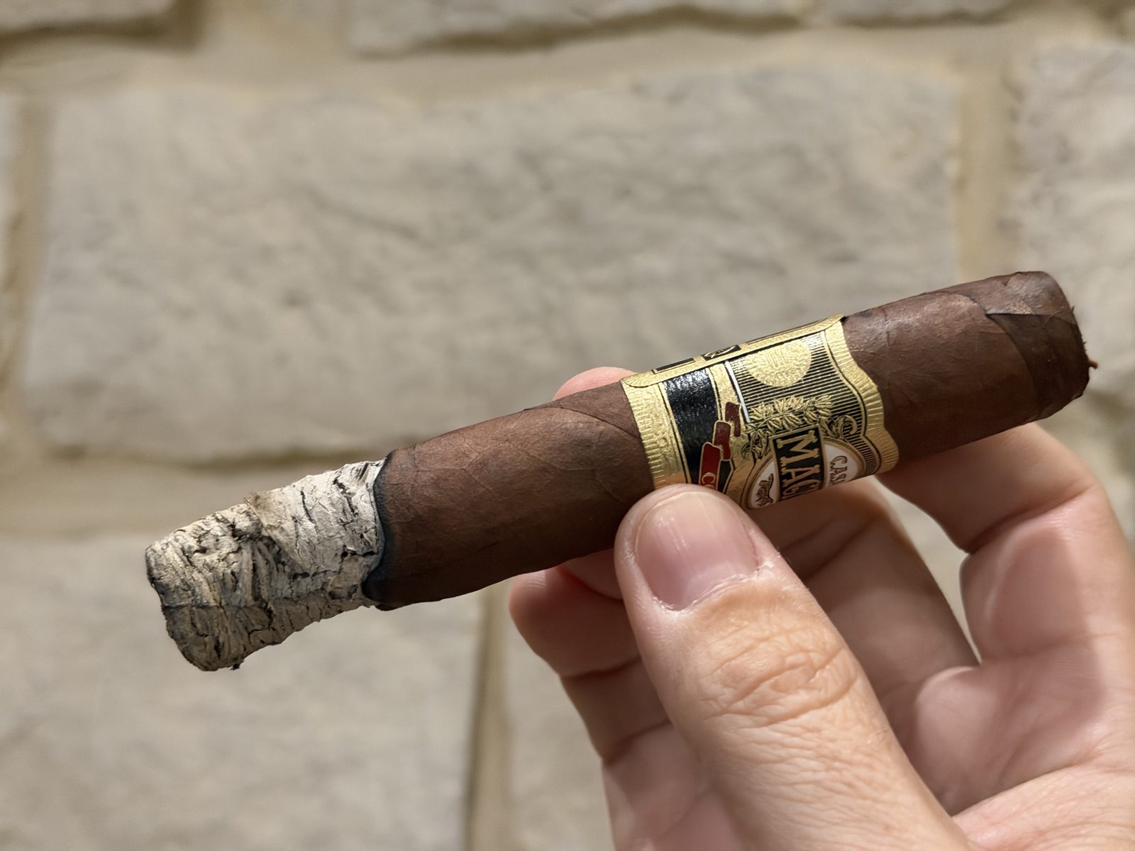 Casa Magna Colorado Robusto at the end of the first third with a long, flaky gray and white ash and an uneven burn line, held in hand against a stone wall