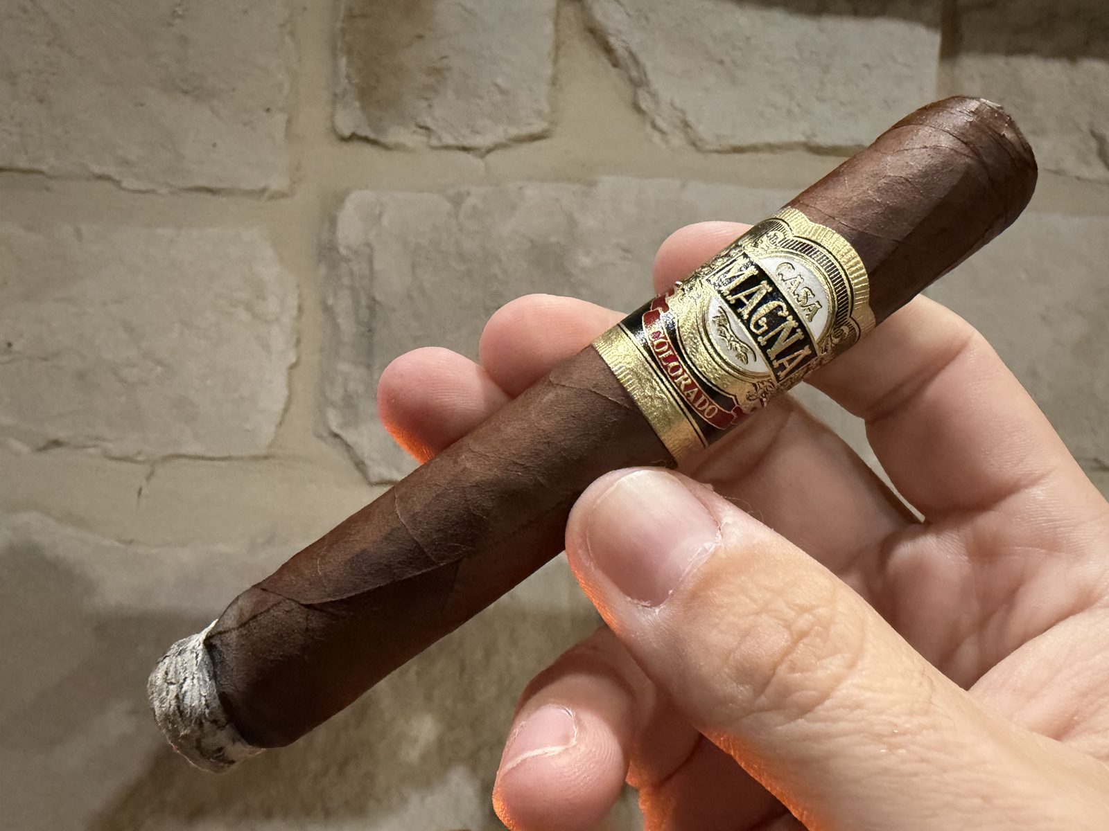 Casa Magna Colorado Robusto held in hand against a stone wall, medium-dark brown wrapper with prominent veins, gold, black and red Casa Magna Colorado band, about a quarter inch burned