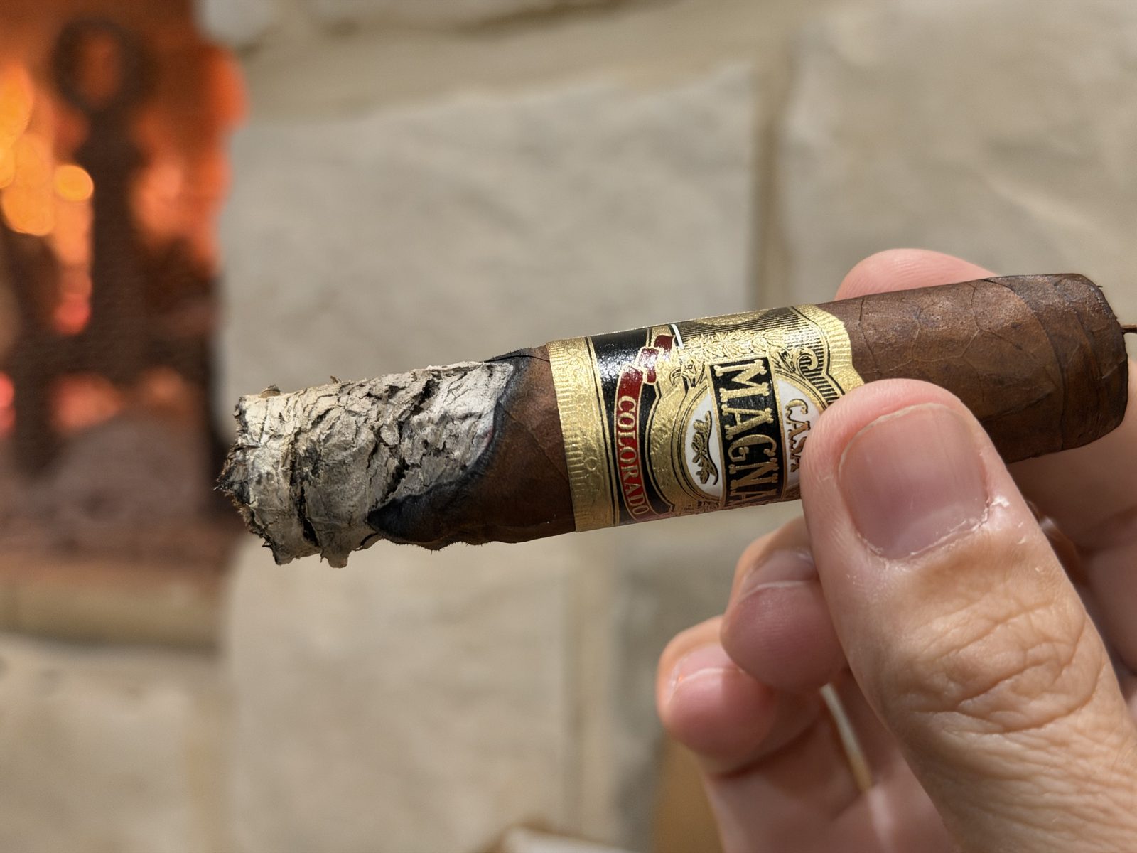 Casa Magna Colorado Robusto late in the second third, the burn line nearly touching the band on one side while the other side runs far behind, fire glowing in a stone fireplace in the background