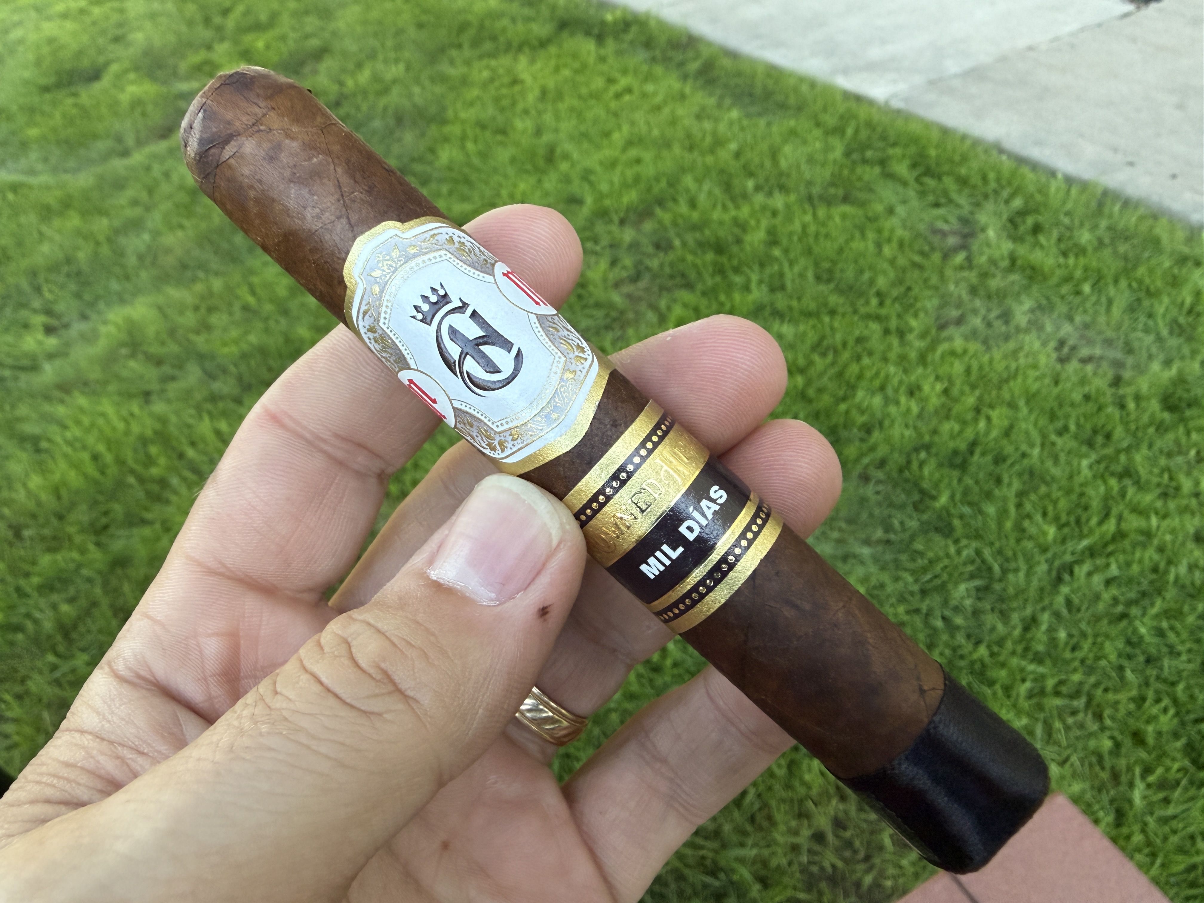 Unlit Crowned Heads Mil Días Maduro Edmundo held over grass, dark chocolate-brown Connecticut Broadleaf wrapper with the ornate white-and-gold crown band and the black MIL DÍAS secondary band