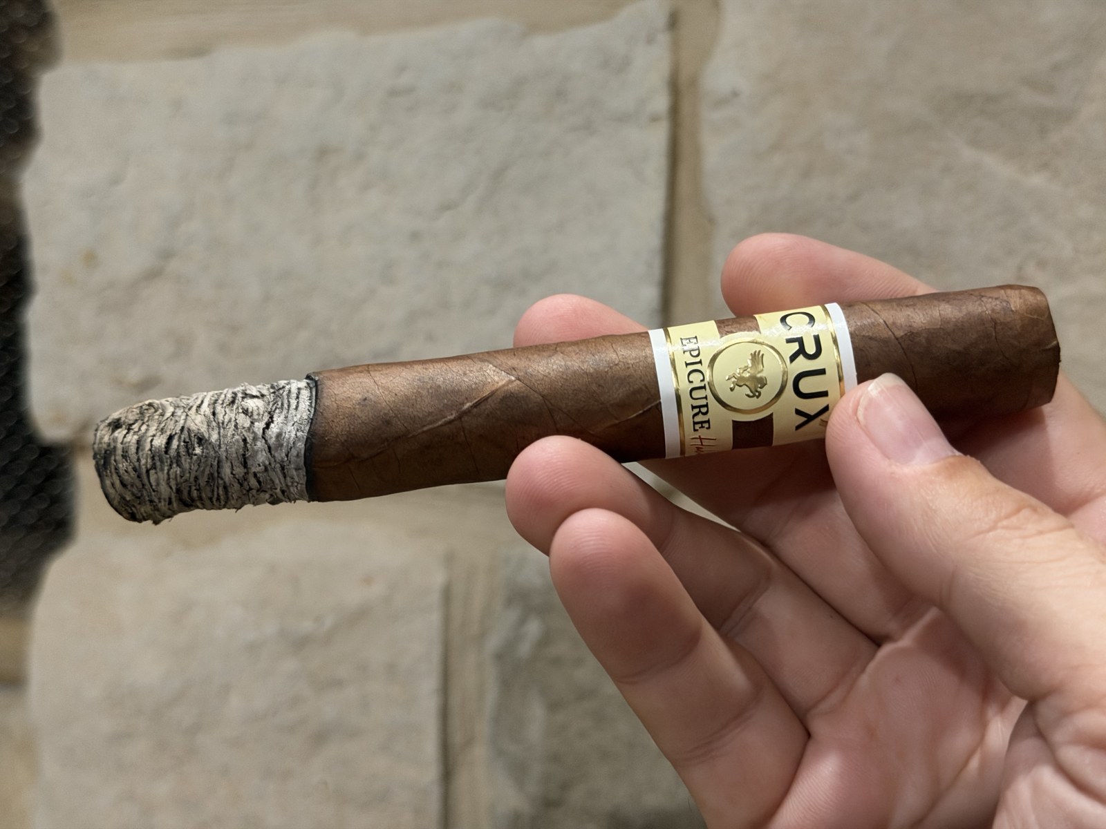 Crux Epicure Habano Toro in the first third with a tight light gray ash stack about an inch long, held against a stone wall