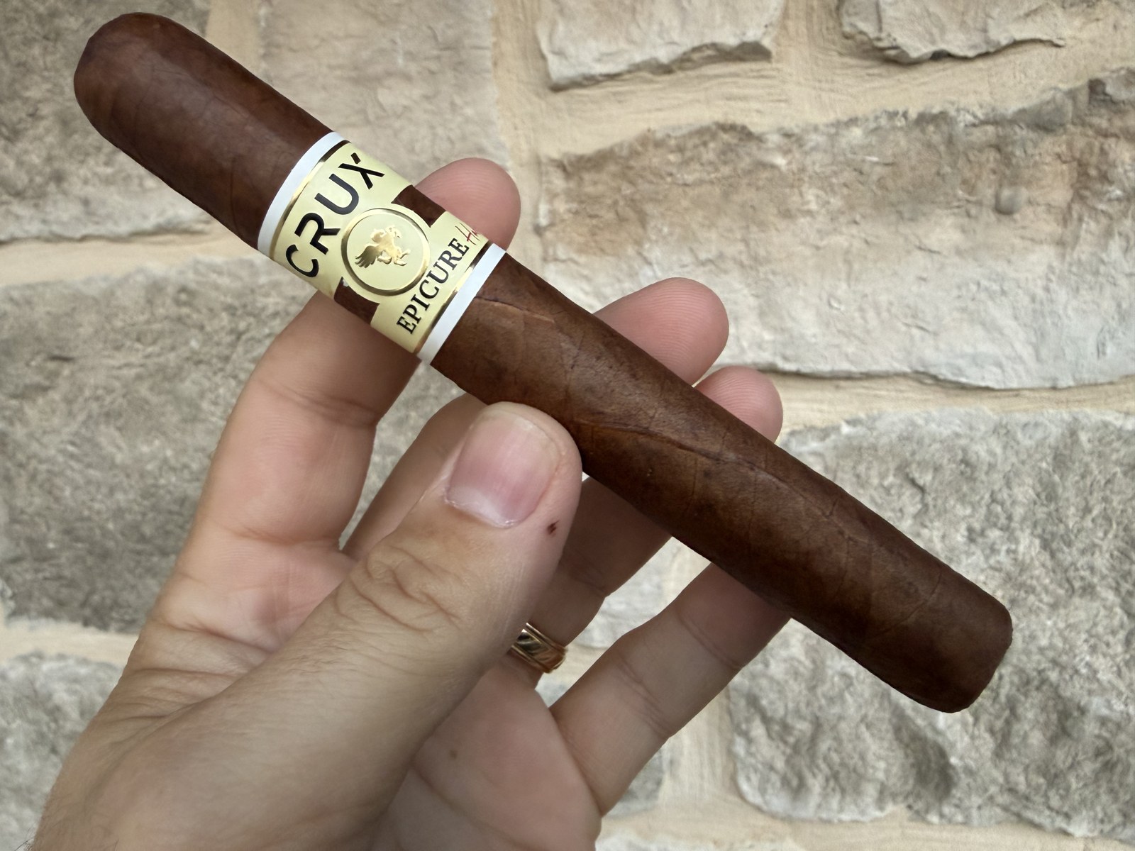 Unlit Crux Epicure Habano Toro held in hand against a stone wall, reddish dark brown wrapper with the cream and gold Crux Epicure Habano band