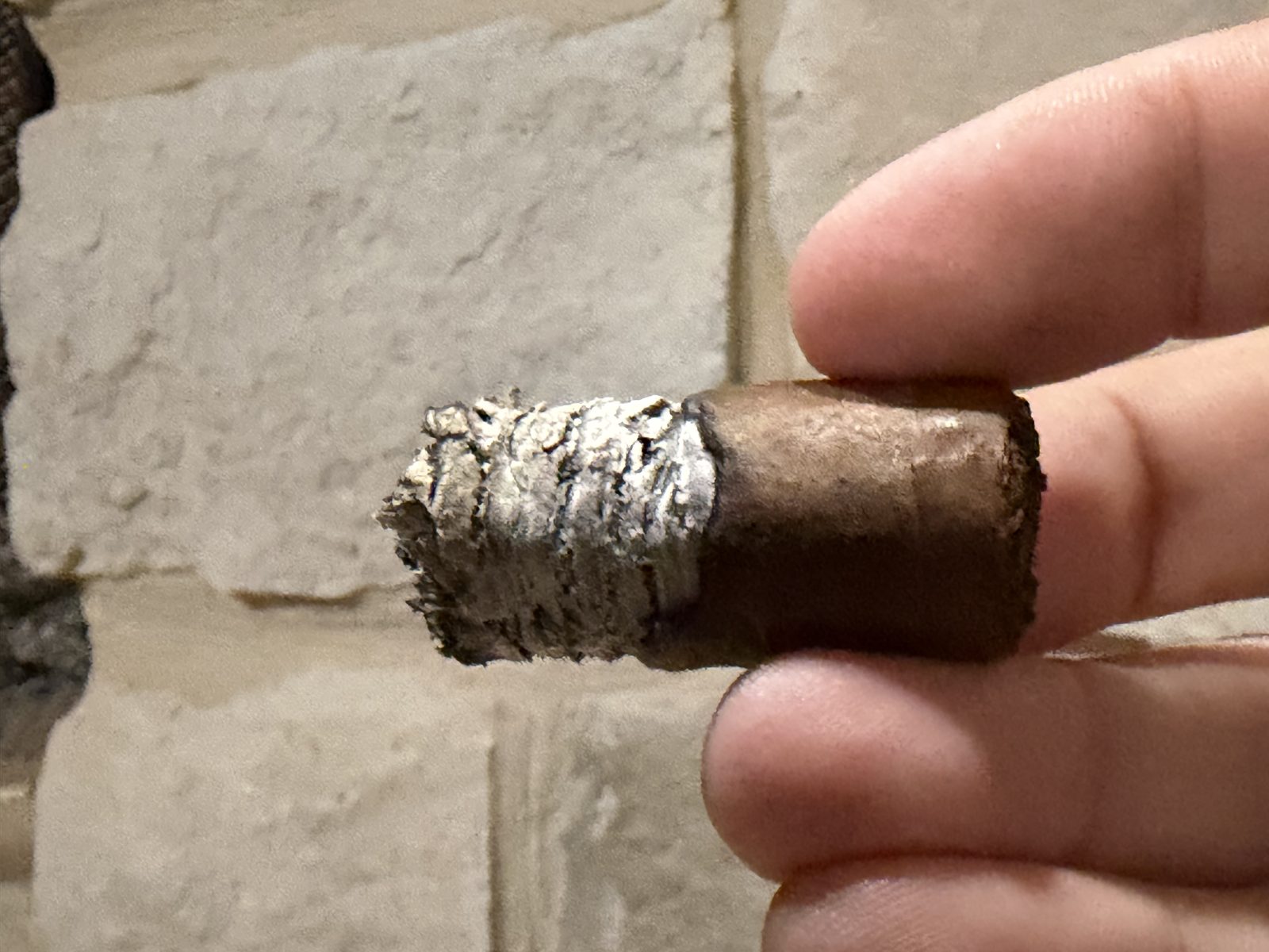 Nub of the Diesel Uncaged Toro held between fingers, a short ragged gray ash on a light brown wrapper with no band, in front of a stone wall
