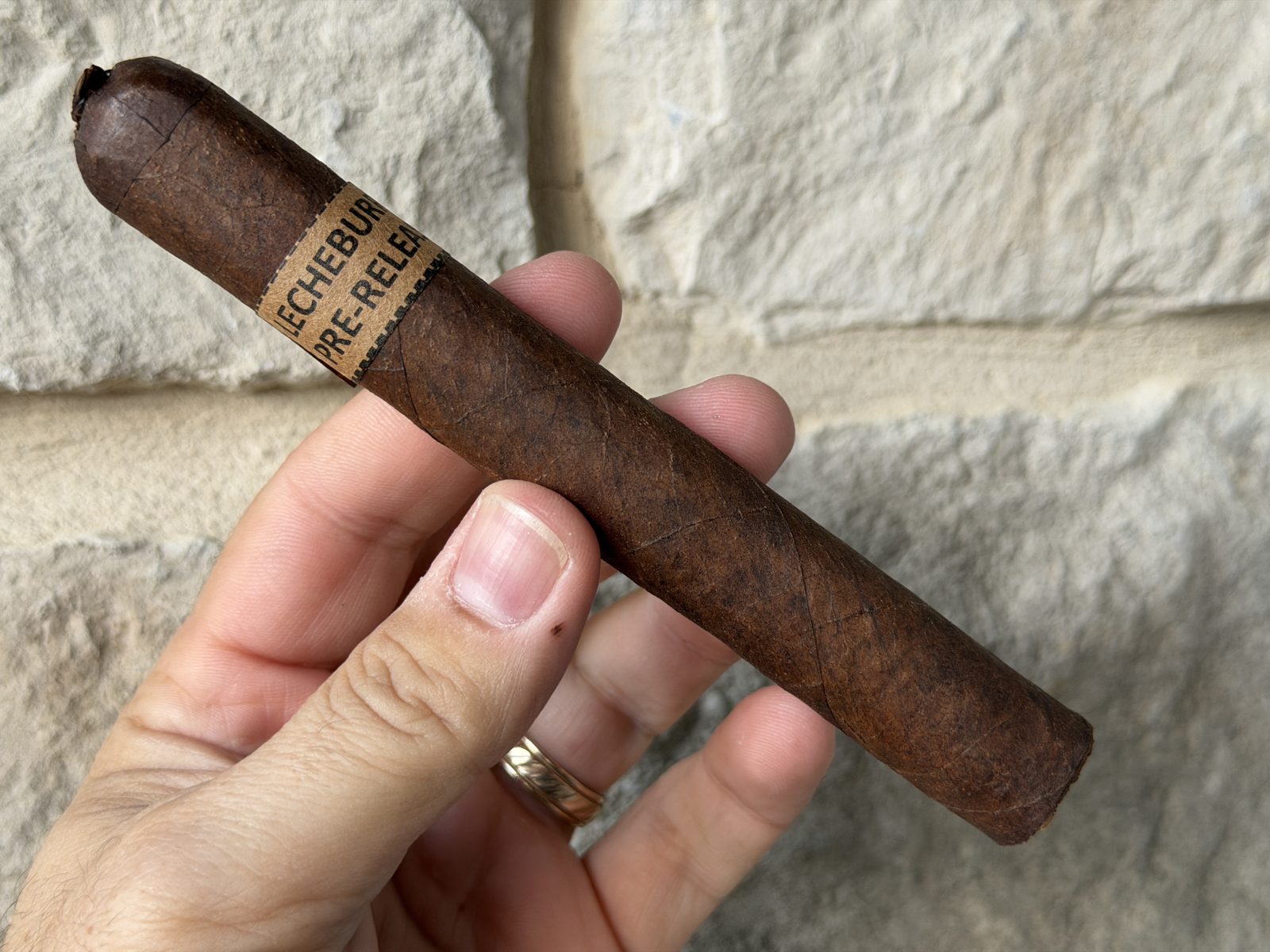 Unlit Dunbarton LecheBurra pre-release toro held in hand against a stone wall, dark milk-chocolate San Andres wrapper with the tan pre-release band and a pigtail cap