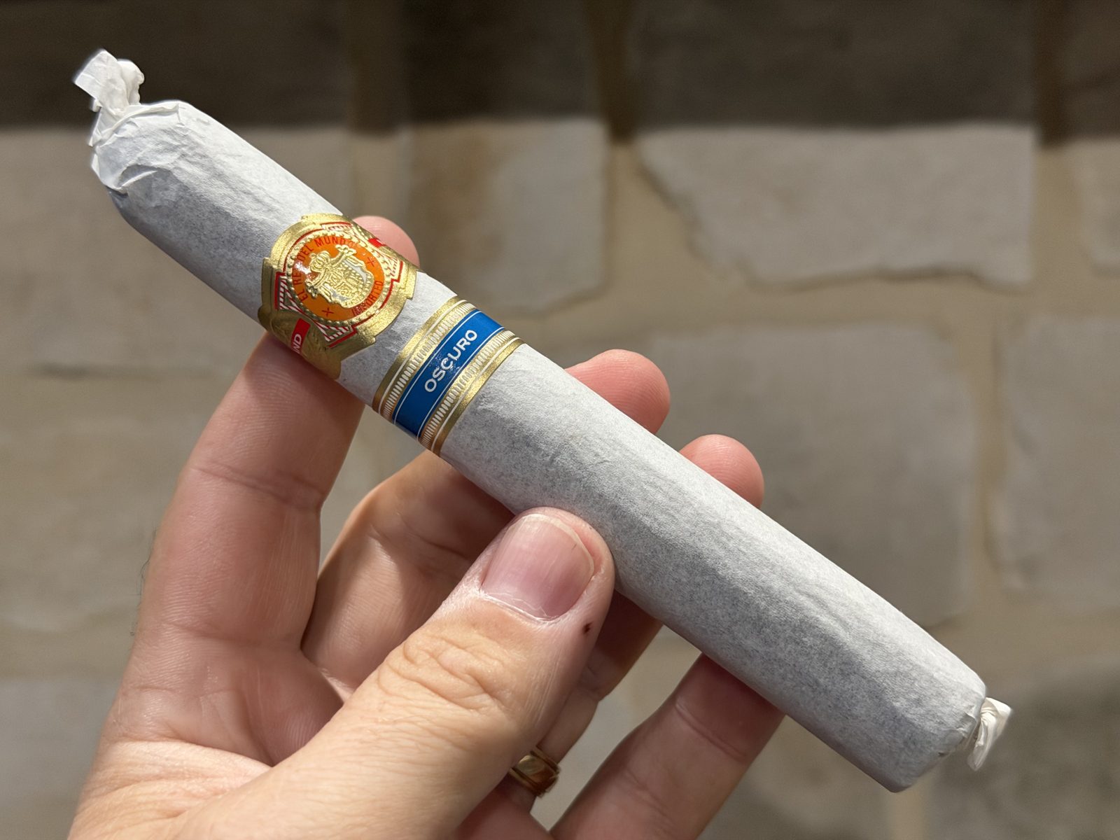 El Rey del Mundo Oscuro Robusto Larga still wrapped in its white tissue paper, held in hand against a stone wall, with the orange and gold El Rey del Mundo band and the blue Oscuro secondary band showing through