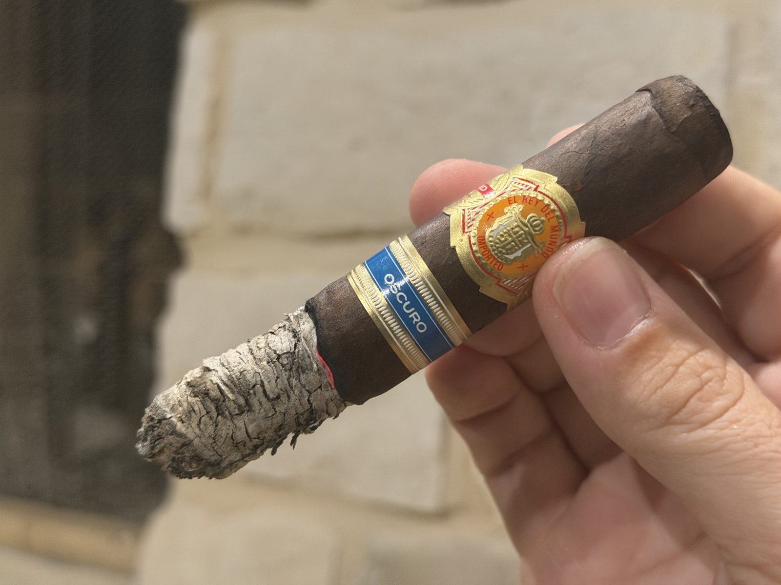 El Rey del Mundo Oscuro Robusto Larga at the end of the second third with a gray ash stack and a chunk of ash sticking out one side like a wing, held in hand in front of a stone fireplace