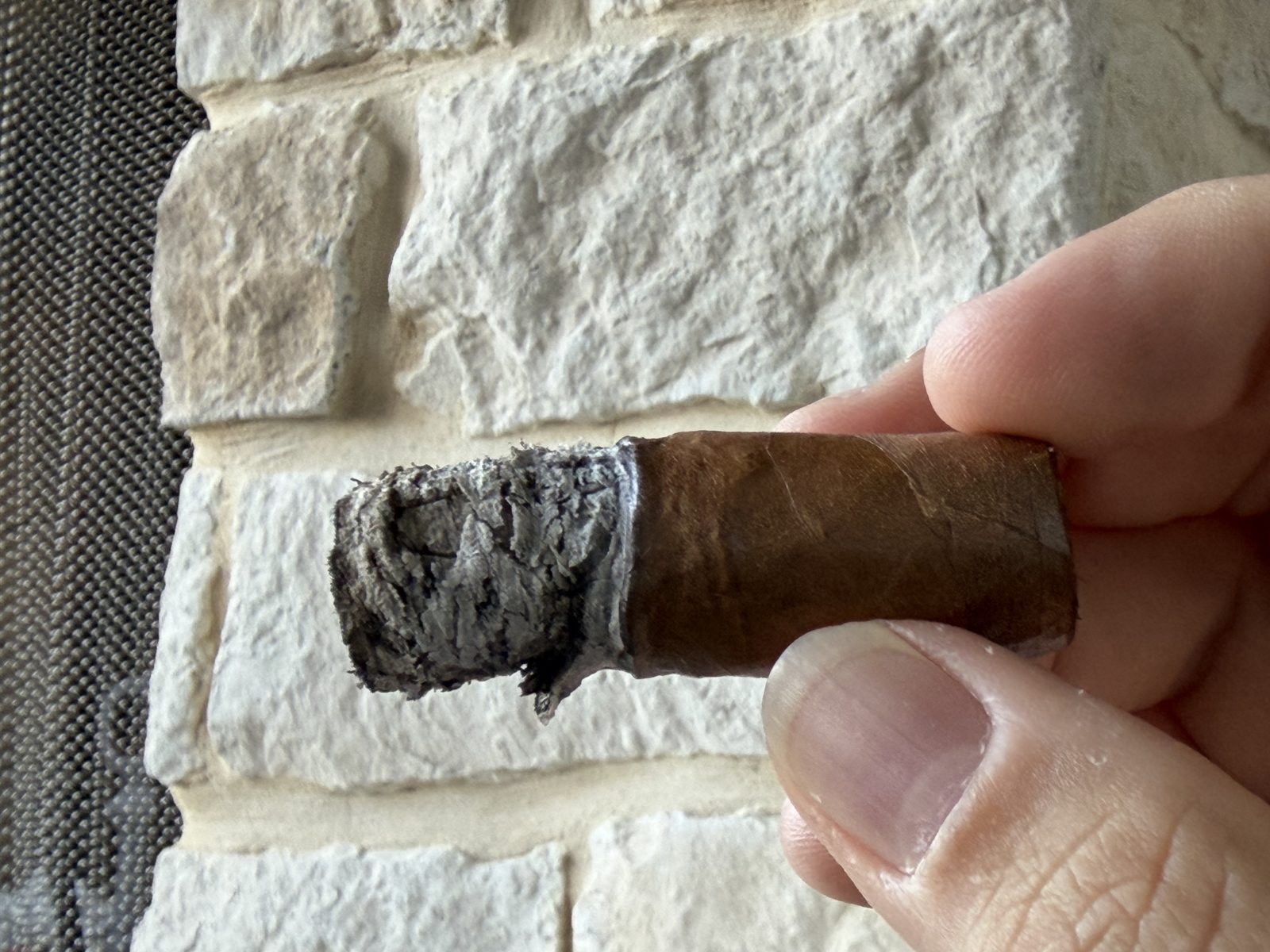Nub of the Fuente Fuente Opus X Perfecxion X held between fingers, band removed, with an uneven burn and a long unburnt finger of wrapper