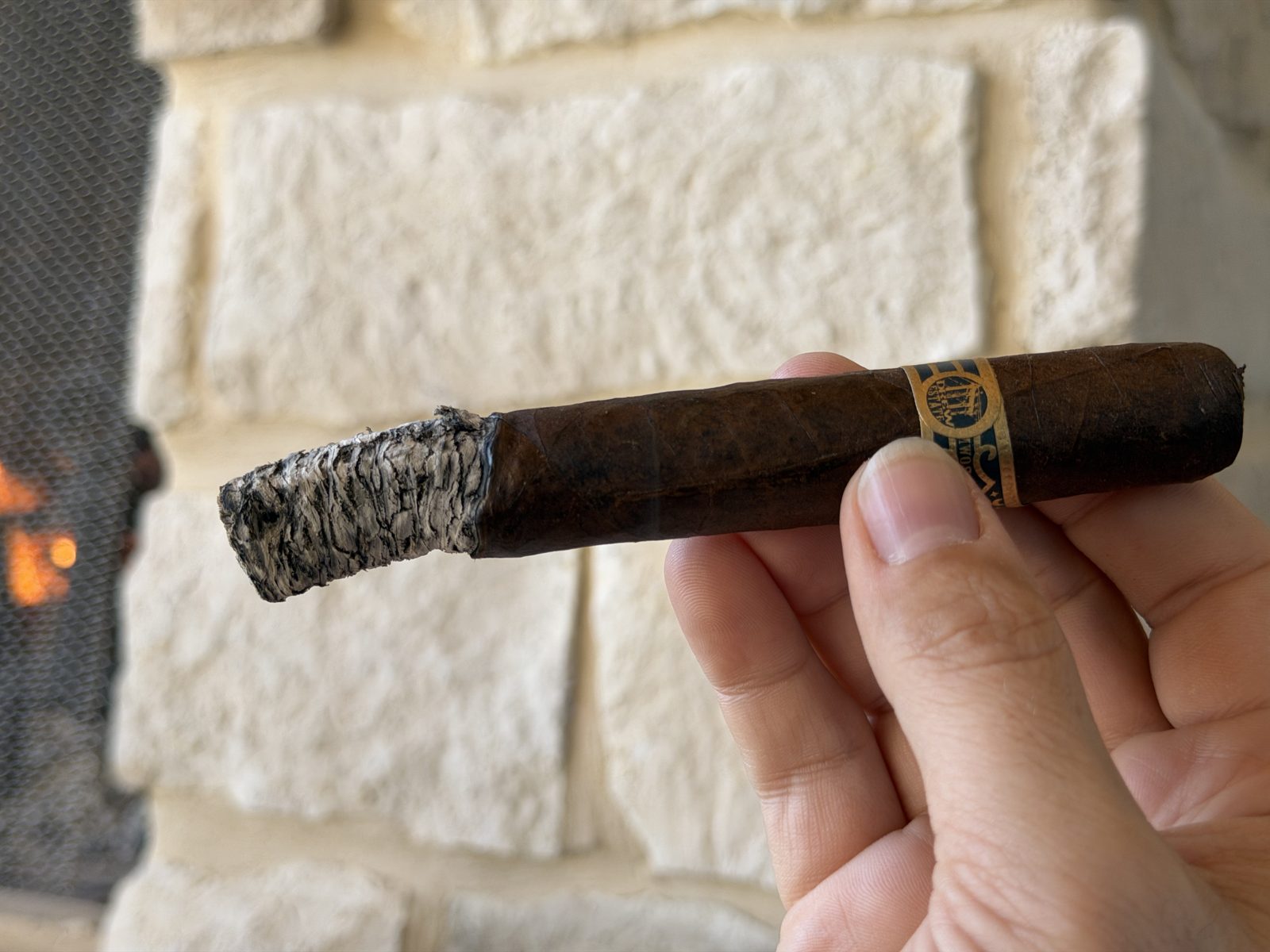 Liga Undercrown Maduro Gran Toro at the end of the first third with about an inch of tight, layered gray and white ash, held in hand in front of a stone outdoor fireplace with a fire going