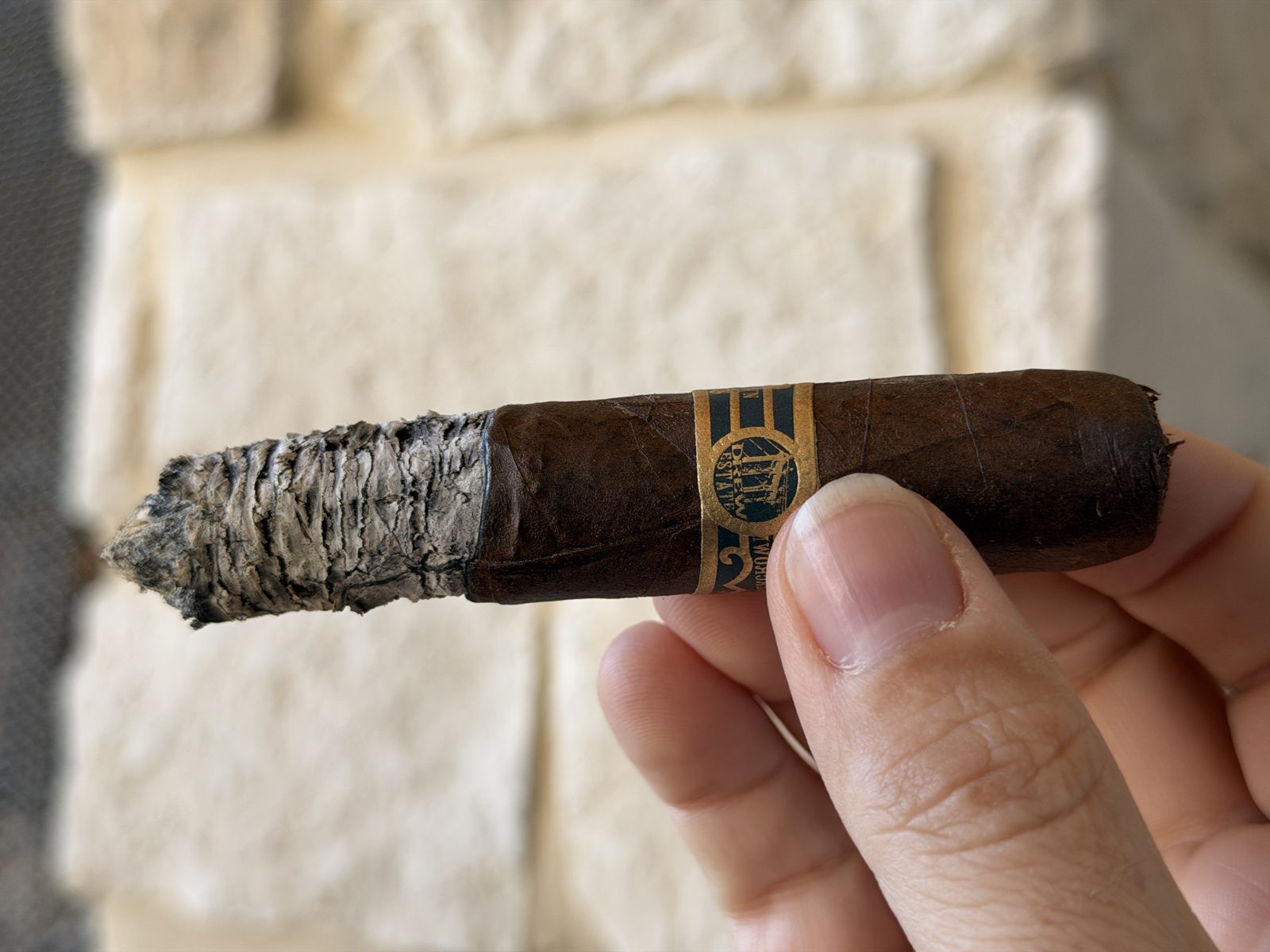 Liga Undercrown Maduro Gran Toro at the end of the second third with a long, layered gray ash and a slightly wavy burn, held in hand against a stone wall