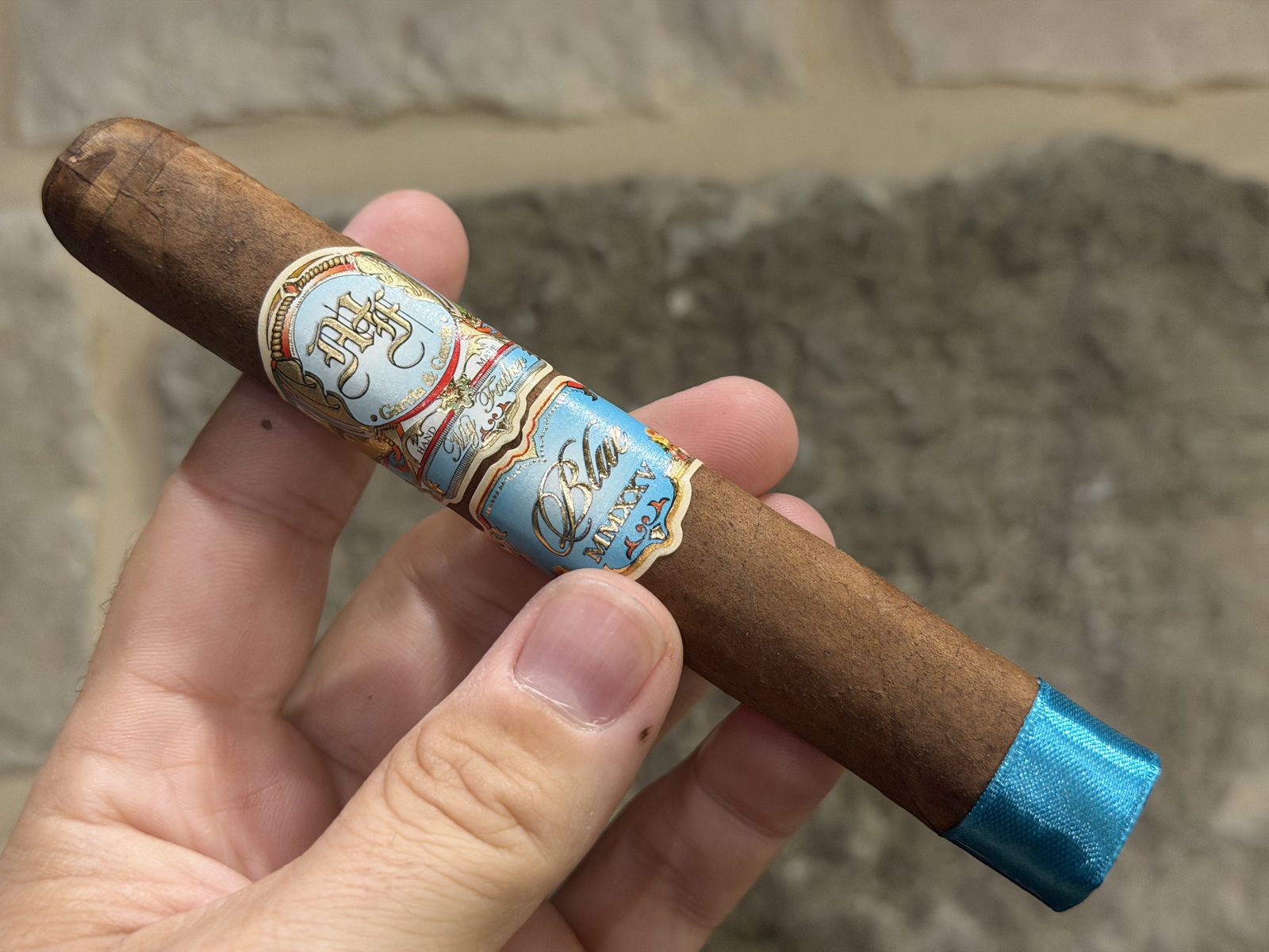 Unlit My Father Blue Toro held in hand, medium brown Connecticut Broadleaf Rosado wrapper with the My Father main band, light blue Blue secondary band, and a light blue foot band