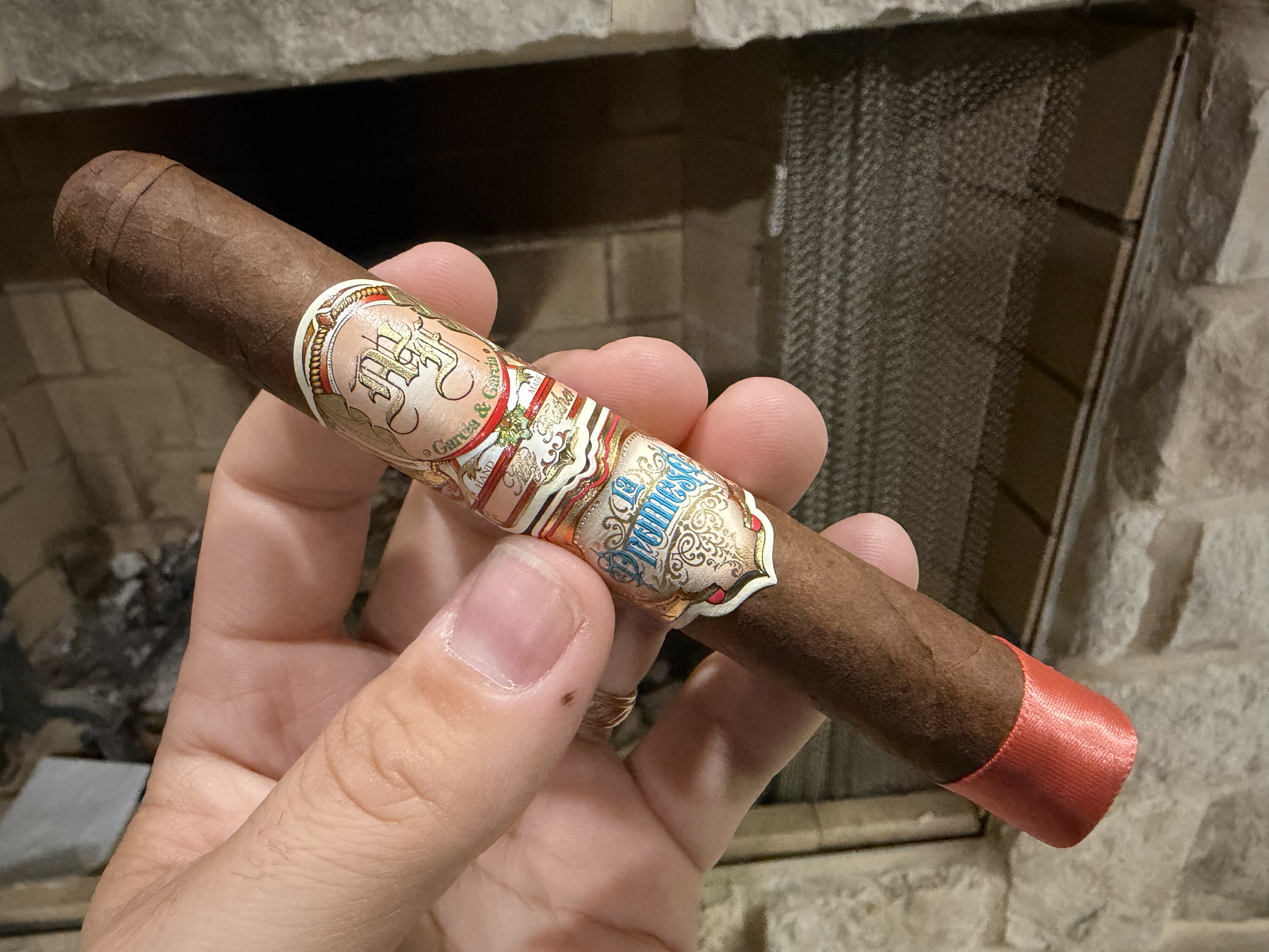 Unlit My Father La Promesa Corona Gorda held in hand in front of a stone fireplace, dark brown Habano Rosado Oscuro wrapper with the My Father main band, blue La Promesa second band, and reddish-pink foot ribbon