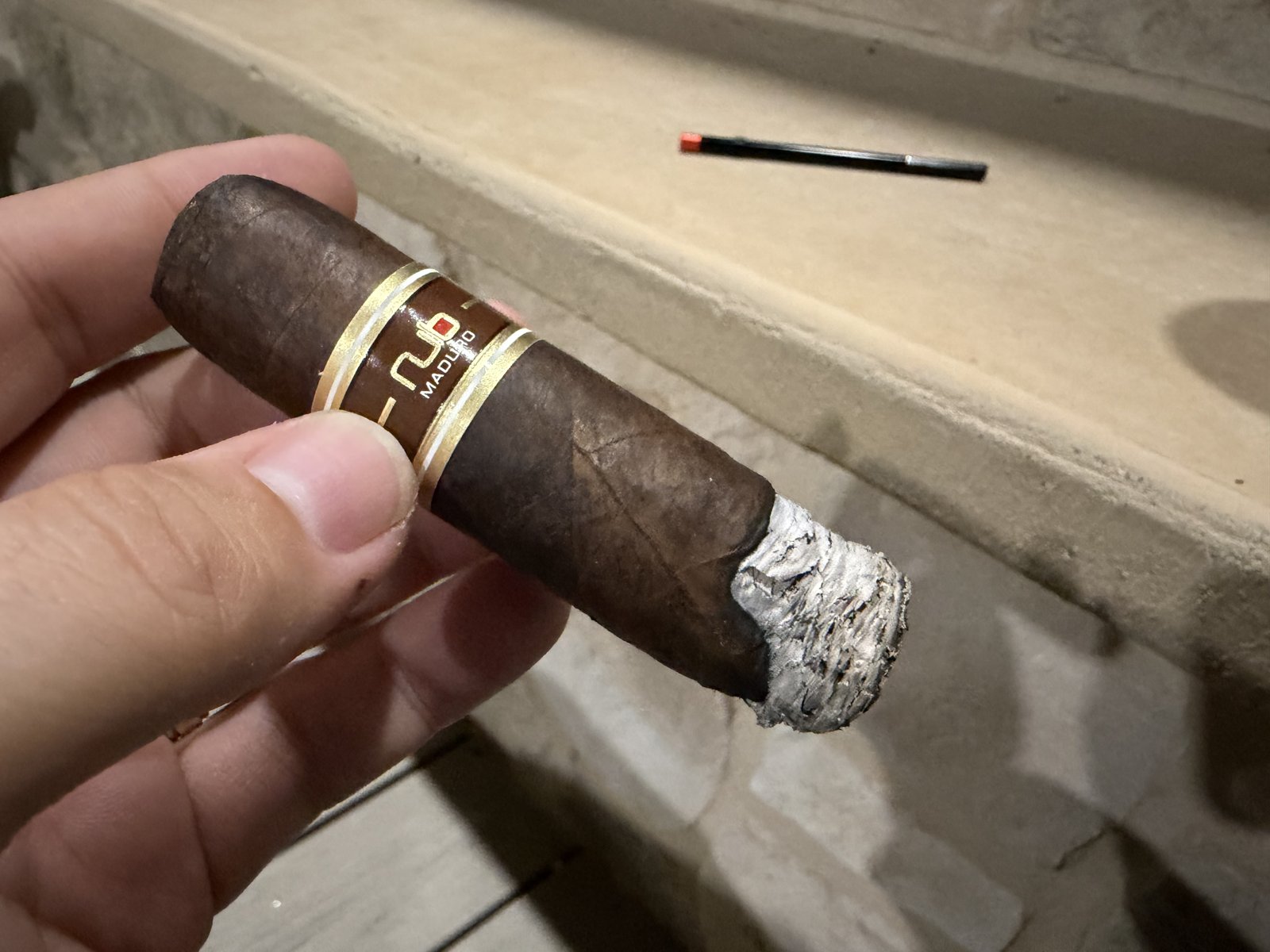 Nub Maduro 460 lit and held in hand, ash forming with one long finger of wrapper lagging on one side