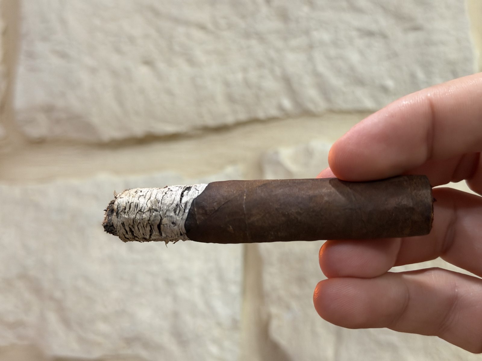 Oliva Master Blends 3 Churchill in the final third with the band removed, short white ash on the foot, held in fingertips against a stone wall