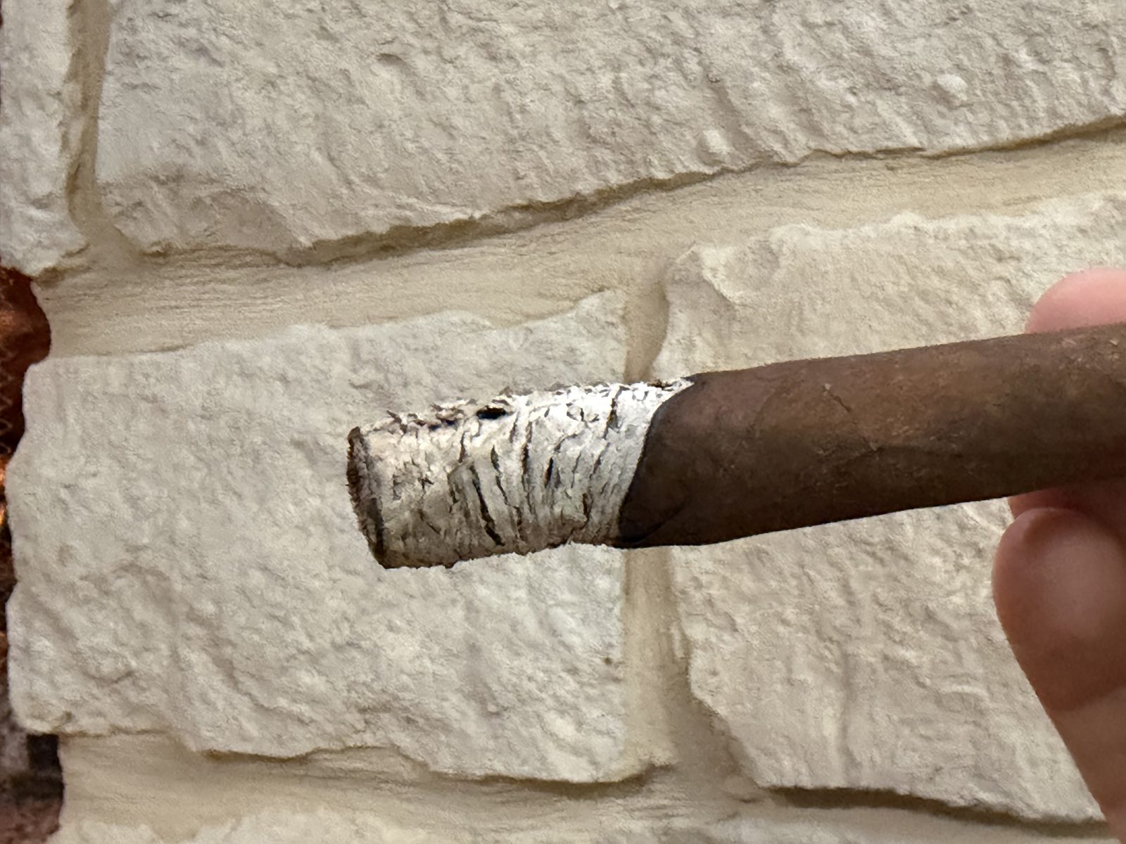 Oliva Master Blends 3 Churchill about an inch in with a compact white ash and a burn line running ahead on the top side, band on, held against a stone wall with the fire in the background