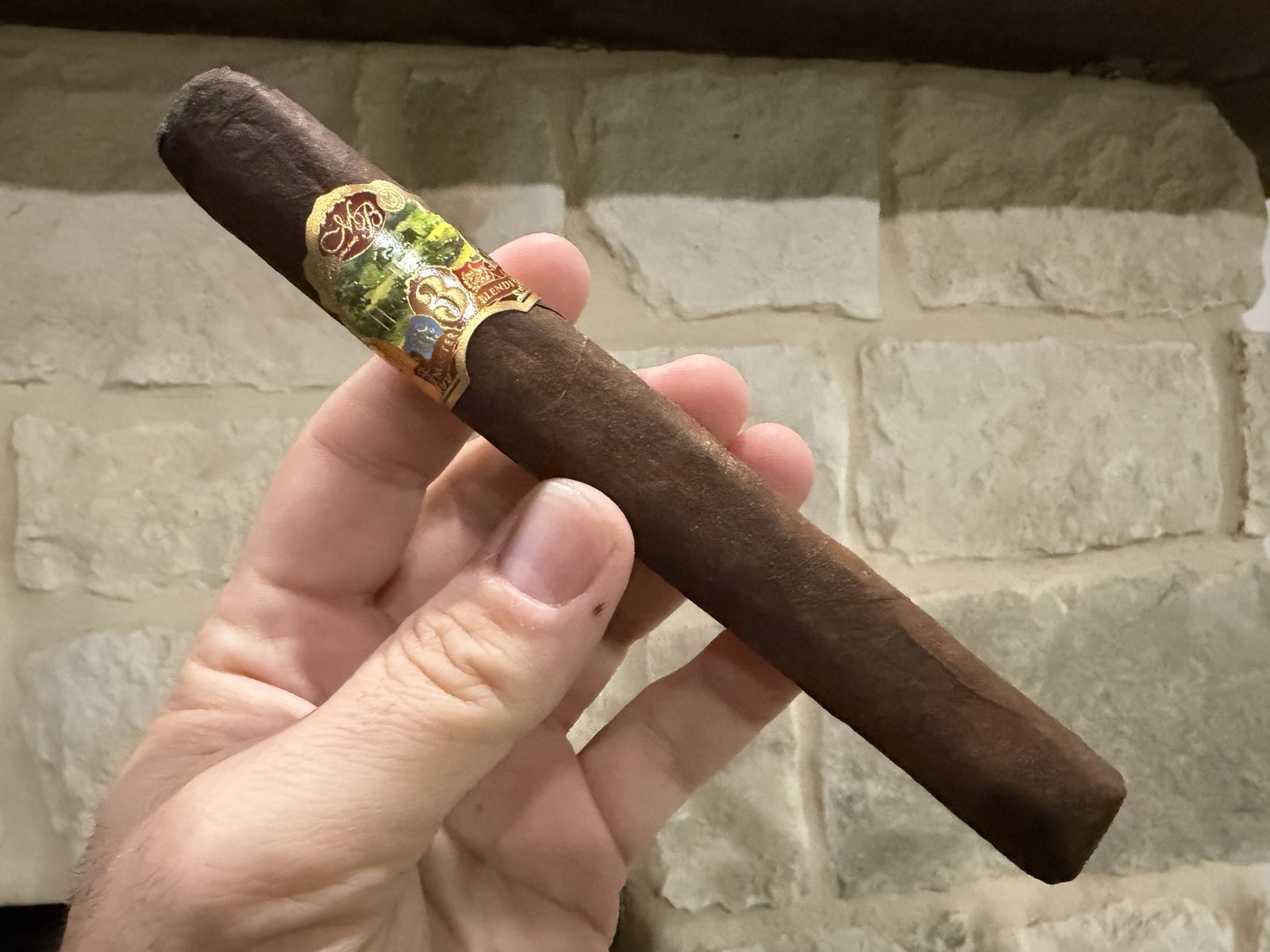 Unlit Oliva Master Blends 3 Churchill held in hand against a stone wall, dark brown smooth wrapper with the gold and green Master Blends 3 band