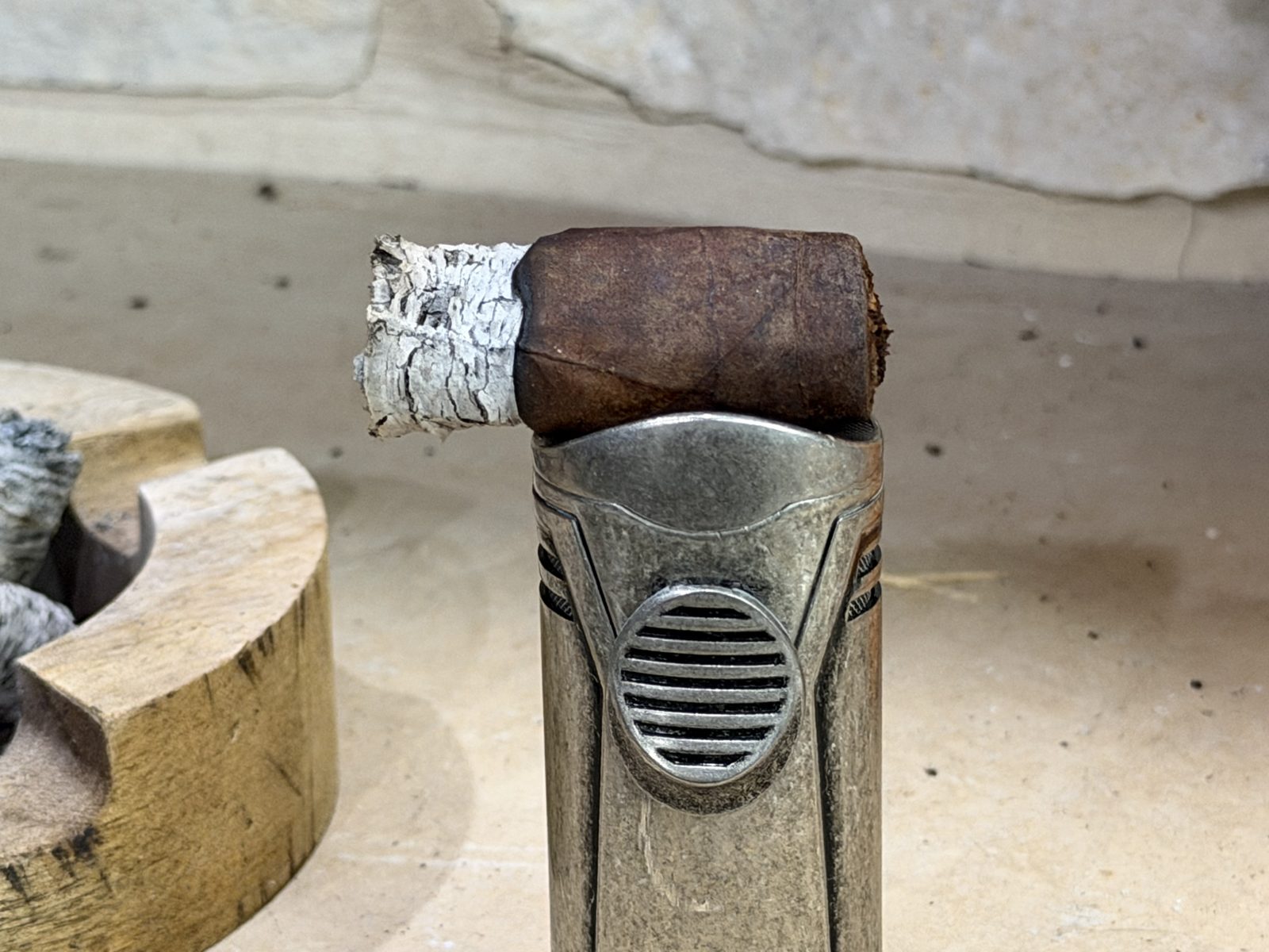 The last half inch of the Oliva Master Blends 3 Churchill resting on top of a torch lighter next to a wooden ashtray, a short white ash on the foot