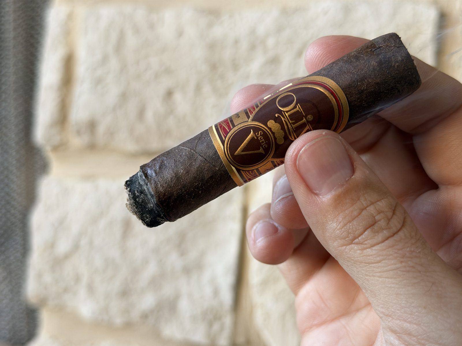 Oliva Serie V Maduro Toro after the first relight about halfway through the second third, foot freshly lit, secondary band removed with only the main Serie V band left, held in hand against a stone wall