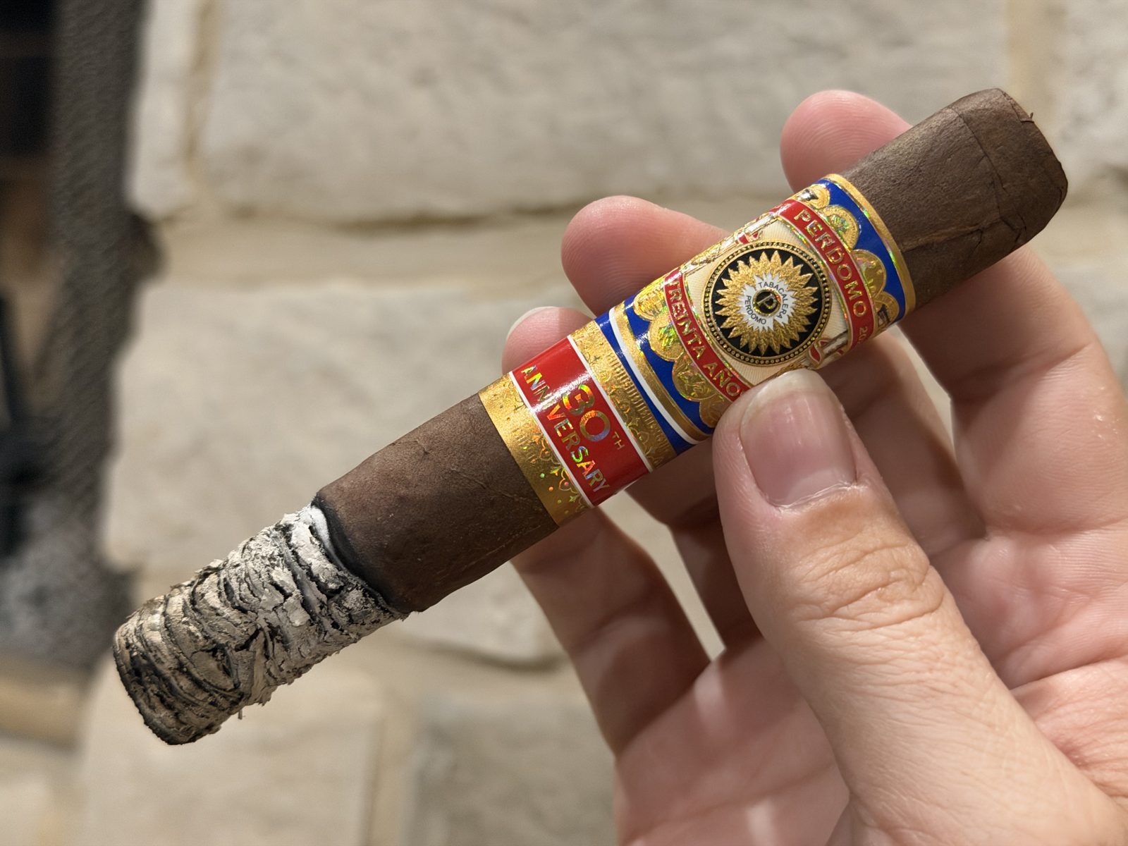 Perdomo 30th Anniversary Maduro Epicure near the end of the first third with about an inch of stacked light gray ash, band still on, held in hand against a stone wall