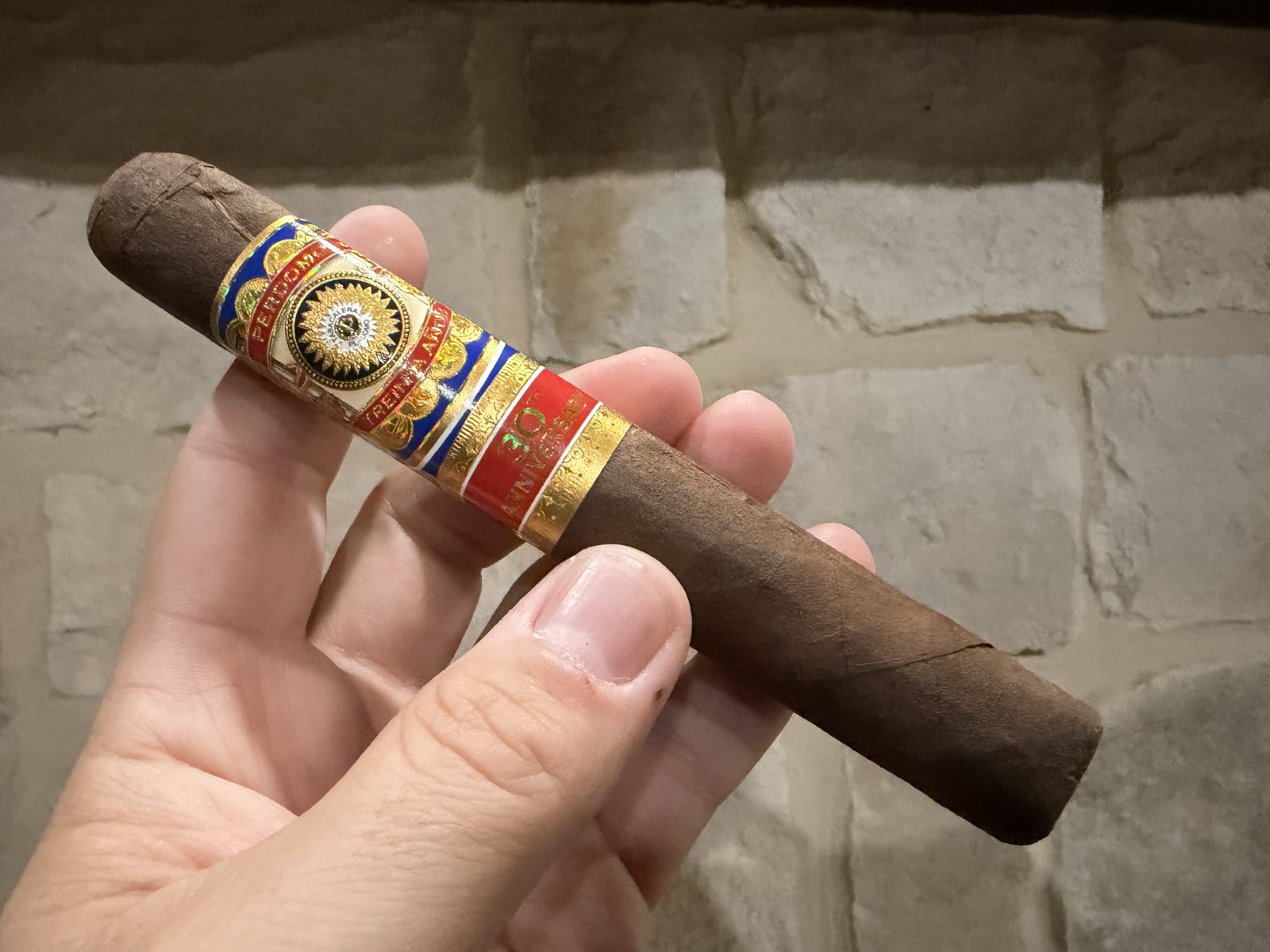 Unlit Perdomo 30th Anniversary Maduro Epicure held in hand against a stone wall, dark brown box-pressed wrapper with the red, gold and blue 30th Anniversary bands