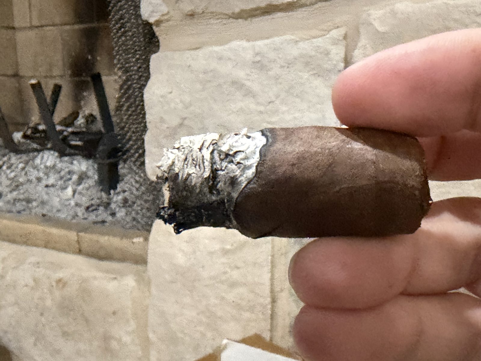 Short stub of the Perdomo 30th Anniversary Maduro Epicure near the end, band off, a crumbling gray ash and a charred, uneven foot, held in fingers in front of a fireplace
