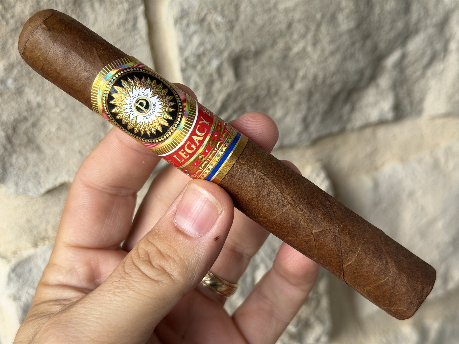 Unlit Perdomo Legacy Nicaraguan Shade-Grown Epicure held in hand against a white stone wall, reddish-brown wrapper with gold and red Legacy band