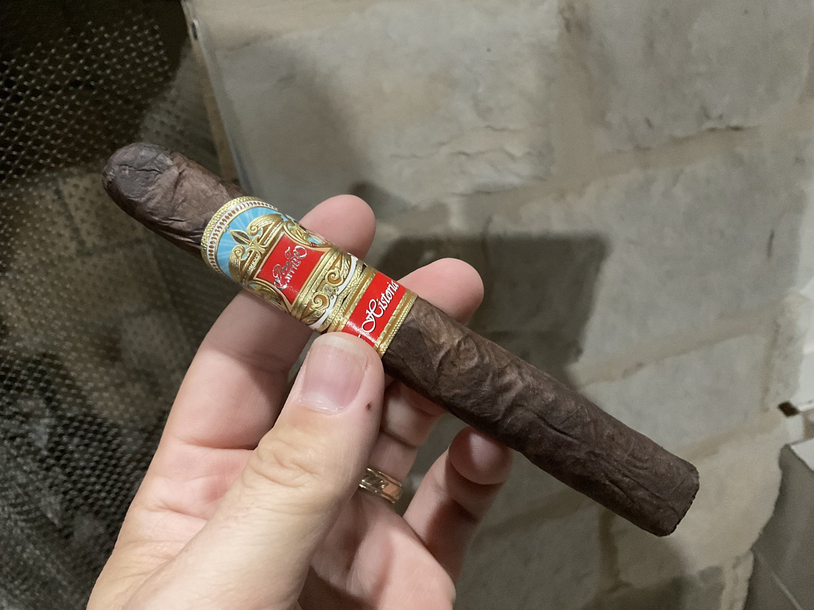 Unlit Perez-Carrillo La Historia Doña Elena held in hand against a stone fireplace, dark wrinkled wrapper with the red and gold La Historia bands and a blue-and-gold ribbon foot band