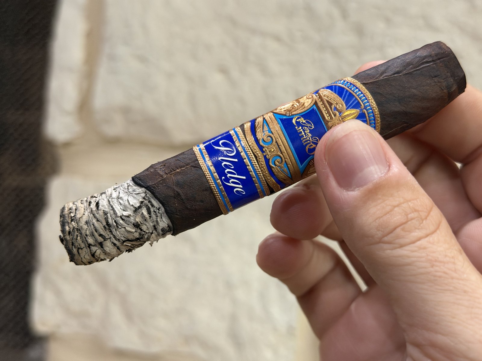 Perez-Carrillo Pledge Prequel lit and held in hand with the first ash still attached, both bands on, white stone wall behind