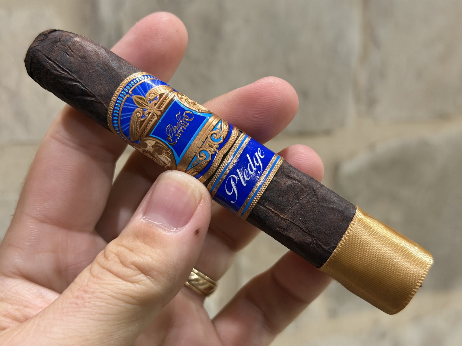 Unlit Perez-Carrillo Pledge Prequel held in hand against a stone wall, very dark wrinkled wrapper with the blue and gold main band, Pledge secondary band, and gold foot ribbon