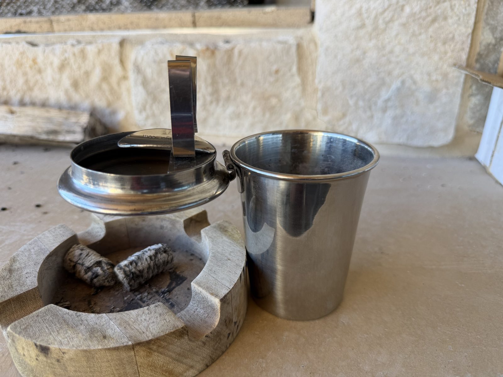 Stinky Cigar Car Ashtray with the lid open, leaning against a wooden ashtray holding cigar ash so it stays upright on a stone ledge