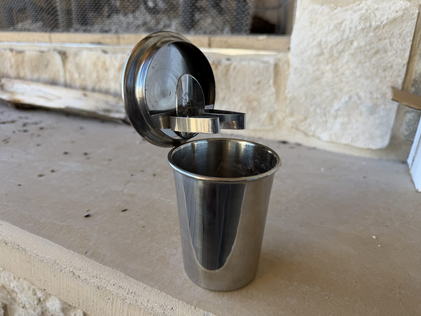 Stinky Cigar Car Ashtray standing on a stone ledge with the hinged lid propped partway open, showing the two stainless cigar wings on the inside of the lid