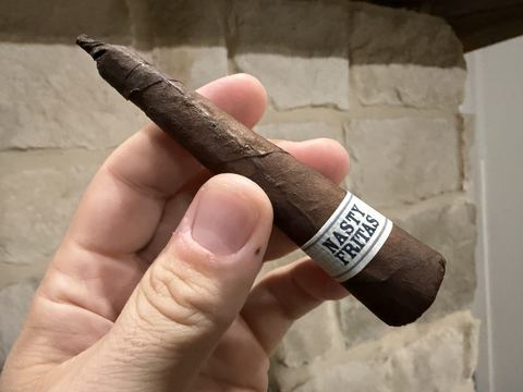 Liga Privada Unico Serie Nasty Fritas held in hand against a stone wall, very dark brown wrapper tapering to a twisted pigtail cap, white Nasty Fritas band near the wide closed foot