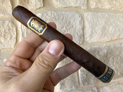 Unlit Liga Undercrown Maduro Gran Toro held in hand against a stone wall, dark brown rustic wrapper with prominent veins, the gold and teal Undercrown band near the head and the teal Maduro foot band